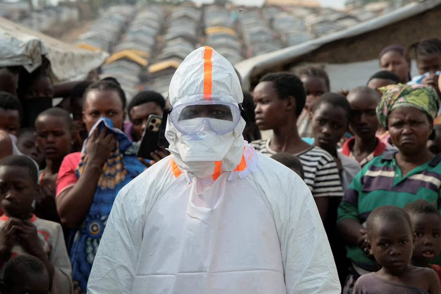 A health worker in personal protective equipment (PPE) stands near displaced people waiting for the burial of suspected Ebola victims at the Kigonze displaced persons camp, one month after an outbreak was declared, in Bunia, eastern Democratic Republic of Congo, June 18, 2026. Gradel Muyisa Mumbere/Reuters