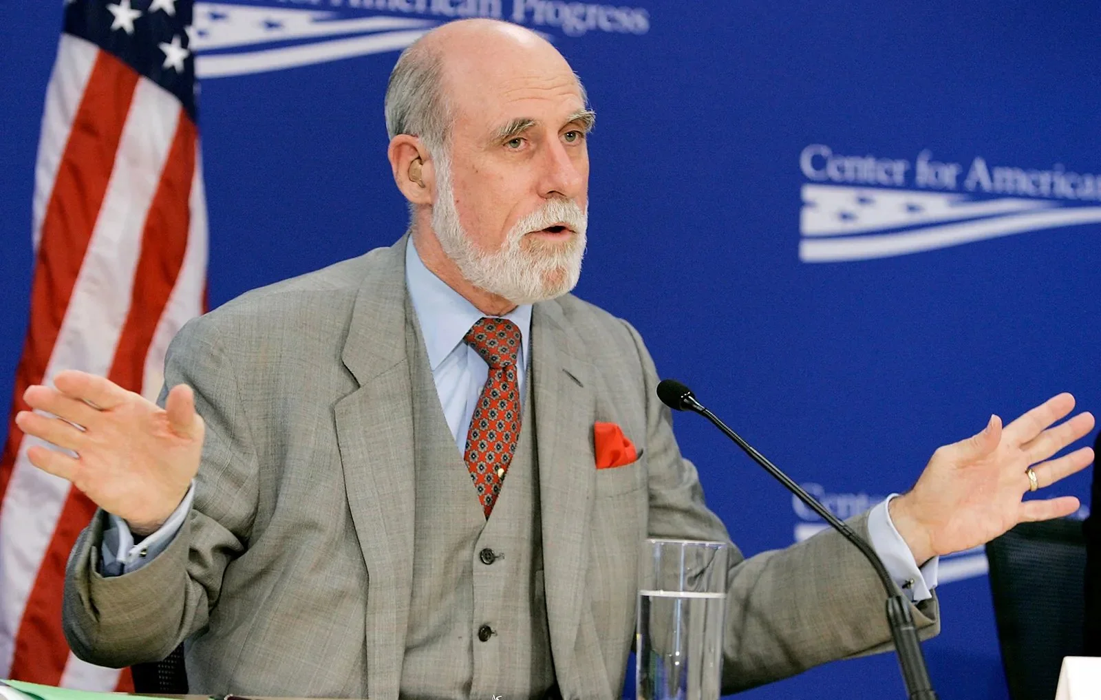 Vinton Cerf, wearing a grey three-piece suit and red tie, is raising both hands while speaking.