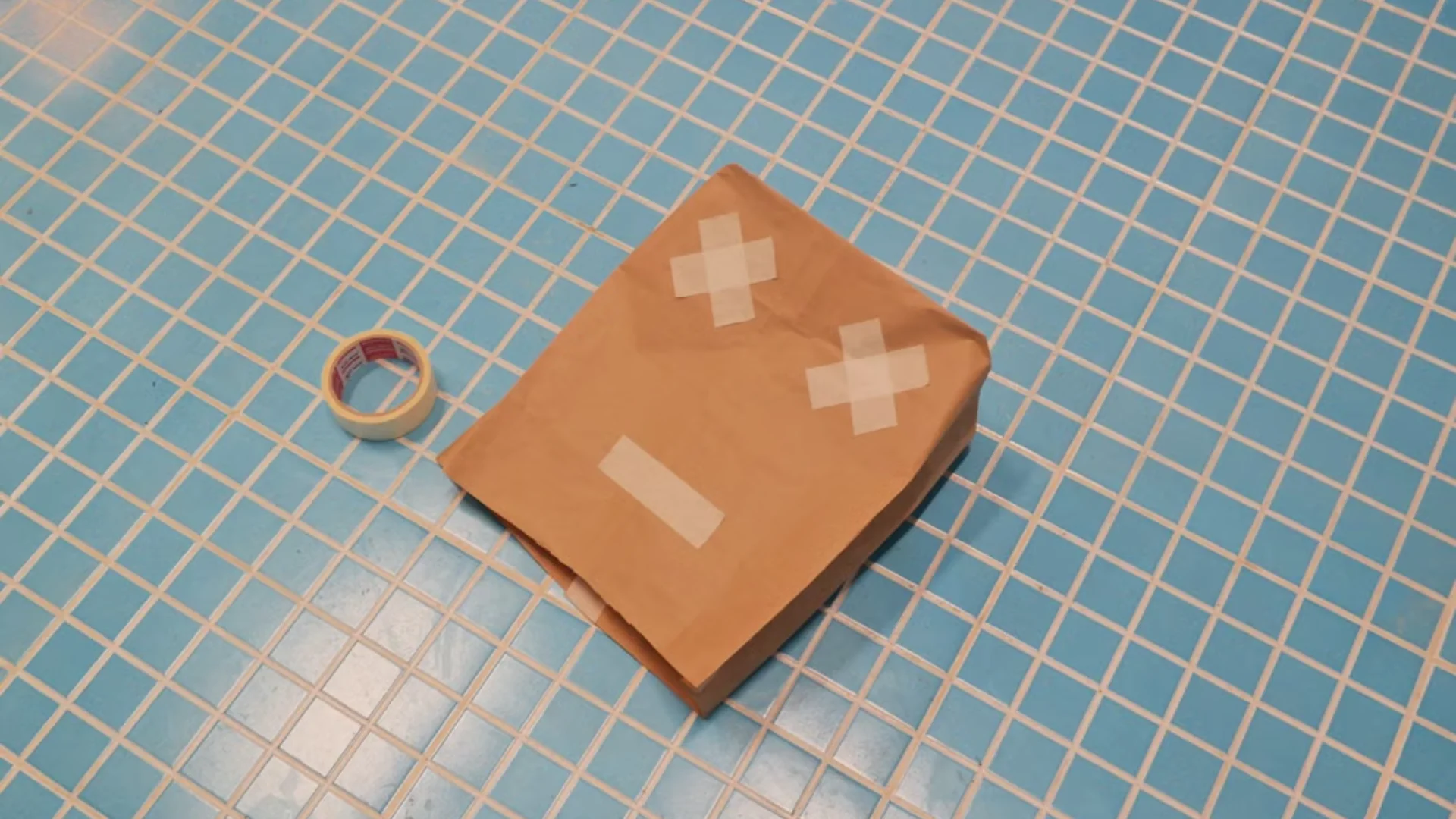 A brown paper bag with an X-eyed tape face on a blue tiled floor. Credit: armağan başaran