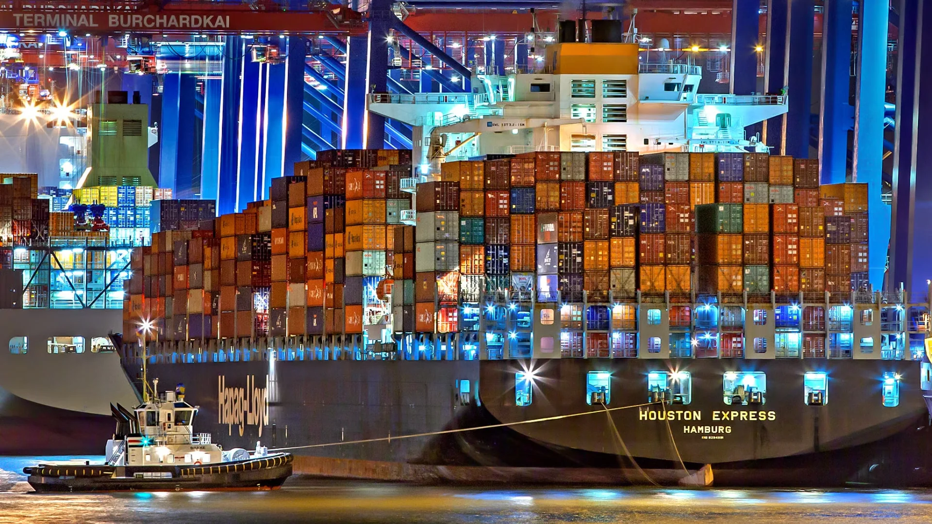 A brightly lit cargo ship at Hamburg port with stacked containers and a tugboat. (Julius Silver)