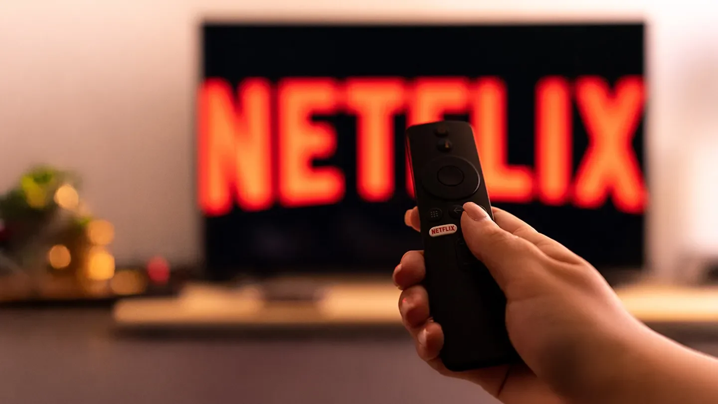 A hand holds a TV remote with a Netflix button in front of a television displaying the company's logo. (Nikos Pekiaridis/NurPhoto via Getty Images / Getty Images)