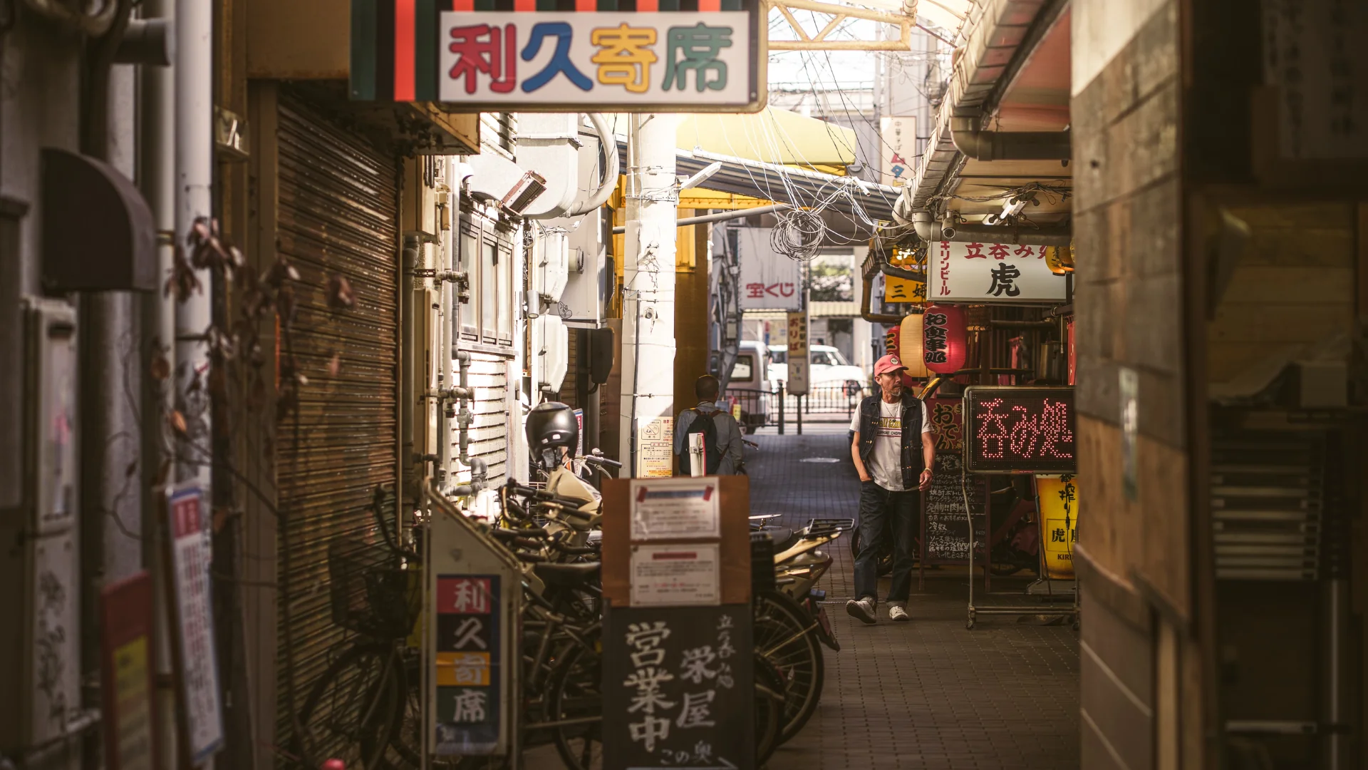 A lively scene of a Japanese alleyway with bicycles, a man in a hat, and bustling small shops. Credit: Paolo Sanchez.