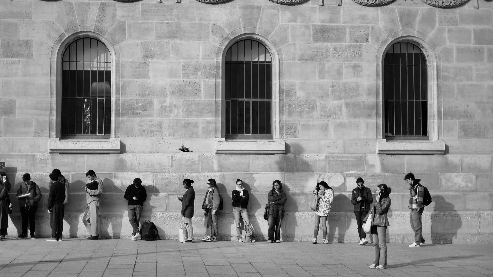 Black-and-white photo of a group of people waiting outside an old stone building with arched windows. Photo: Alina Chern