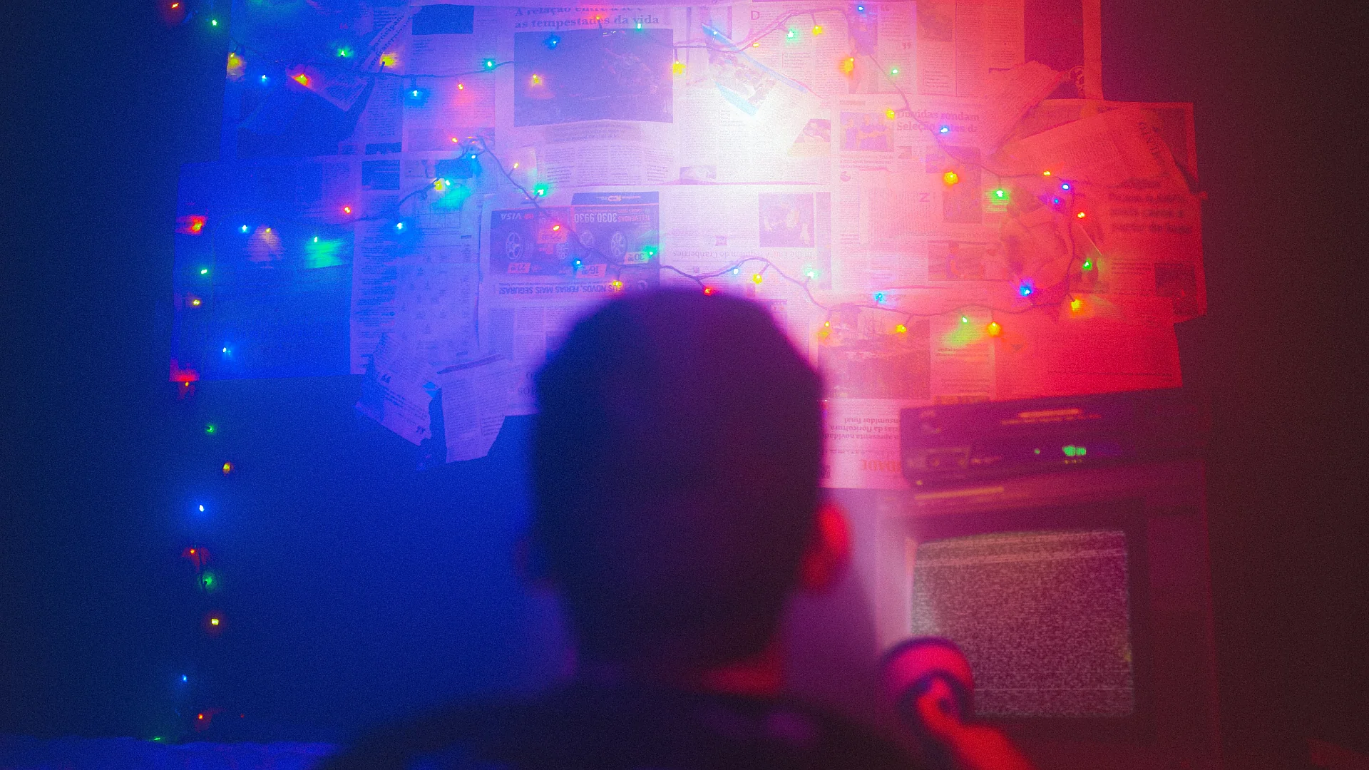 A dark room illuminated by multicolored string lights with a press wall and static on a TV screen. Photo: Matheus Bertel