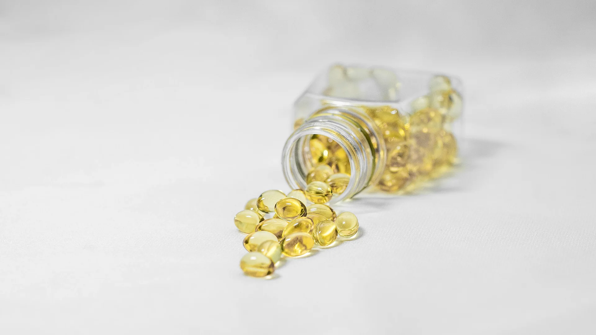 Close-up of yellow softgel capsules spilling out of a clear glass bottle onto a white surface. Photo: Richard Khuptong.